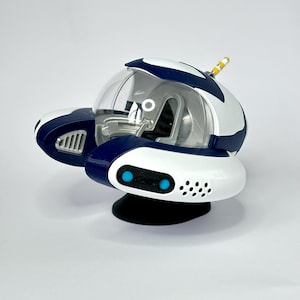 Op de afbeelding: Een wit en blauw speelgoed ruimteschip met een doorzichtige koepel en een zwarte basis. Het ruimteschip heeft twee blauwe ogen en een kleine antenne bovenop.