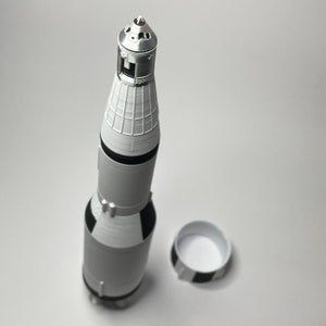K&ouml;nnte beinhalten: Eine wei&szlig;e und schwarze Modellrakete mit silberner Spitze und einem separaten wei&szlig;en und schwarzen Sockel. Die Rakete ist eine Nachbildung einer Saturn V-Rakete.