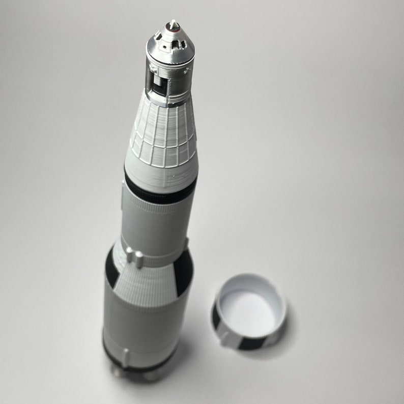 K&ouml;nnte beinhalten: Eine wei&szlig;e und schwarze Modellrakete mit silberner Spitze und einem separaten wei&szlig;en und schwarzen Sockel. Die Rakete ist eine Nachbildung einer Saturn V-Rakete.