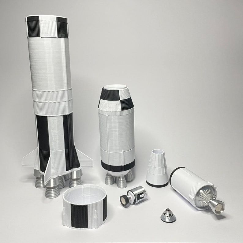 K&ouml;nnte beinhalten: Ein Satz von 3D-gedruckten Modellraketen, darunter eine gro&szlig;e Saturn-V-Rakete, ein kleineres Kommandomodul und eine Mondlandef&auml;hre. Die Raketen sind wei&szlig; mit schwarz-wei&szlig;en Schachbrett-B&auml;ndern.