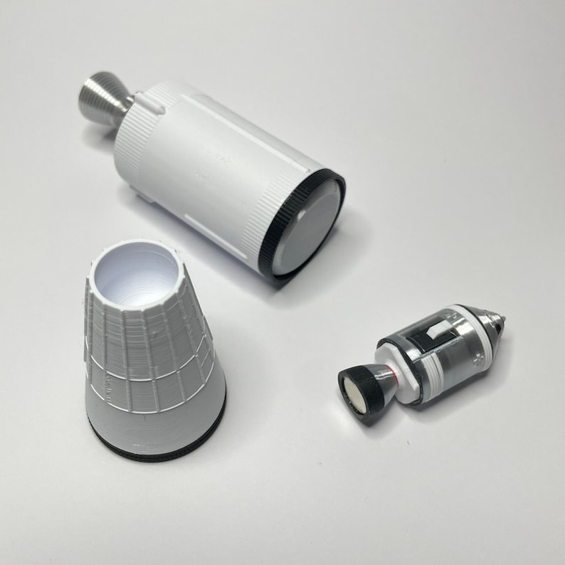 K&ouml;nnte beinhalten: Drei wei&szlig;e Plastikteile f&uuml;r ein Modellraketen-Set. Das gr&ouml;&szlig;te Teil ist ein zylindrischer K&ouml;rper mit einem schwarzen Ring am oberen Ende. Das zweite Teil ist ein kegelf&ouml;rmiger Nasenkonus. Das dritte Teil ist ein kleineres zylindrisches Teil mit einem schwarzen Ring am oberen Ende und einer silbernen Spitze.