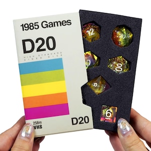 May include: A VHS tape-style box with the text "1985 Games D20 HIGH STANDARD VIDEO DICE 258m VHS D20" printed on the front. The box is open, revealing a set of six iridescent dice with white numbers inside a black foam insert.