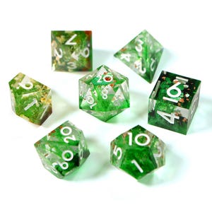 May include: A set of seven polyhedral dice for tabletop games. The dice are clear with a green and gold botanical design inside. Each die has white numbers. The dice are arranged on a white background.