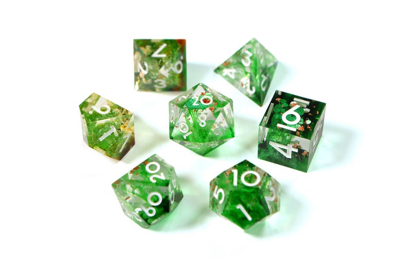 May include: A set of seven polyhedral dice for tabletop games. The dice are clear with a green and gold botanical design inside. Each die has white numbers. The dice are arranged on a white background.