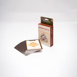 May include: A deck of cards and a box labeled "OBOJIMA SPELLS" with a brown and white color scheme. The box features origami cranes in various colors. A card from the deck is visible, displaying a gold geometric design and the word "OBOJIMA".