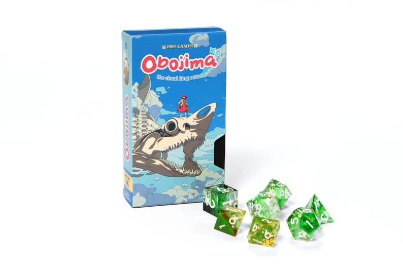 May include: A blue game box titled "Obojima" featuring a shark-like creature illustration. Several green and clear dice with white numbers are scattered in front of the box. The box has a cartoon-style illustration.