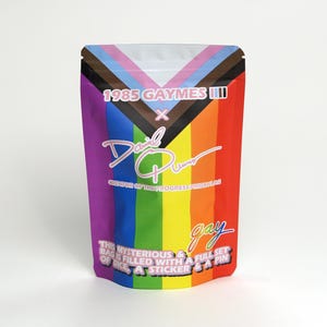 May include: A colorful bag with a rainbow design, featuring text that reads "1985 GAYMES III" and "David Plummer". The bag also includes the word "gay" and the phrase "THE MYSTERIOUS BAG IS FILLED WITH A FULL SET OF DICE, A STICKER & A PIN".