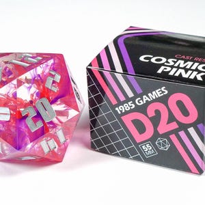 Pode incluir: Um dado de 20 faces rosa e roxo com números prateados. O dado é feito de resina fundida e tem 55 mm de tamanho. O dado é embalado em uma caixa com o texto "1985 Games D20 Cast Resin Cosmic Pink" impresso nela.