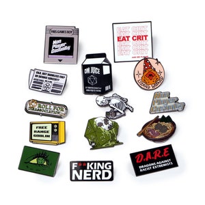 Puede incluir: Una colección de pines esmaltados con varios diseños y texto. Los pines incluyen una consola de juegos retro, un cartón de leche, un esqueleto con una cámara, un dado y frases como "Eat Crit" y "F**king Nerd".