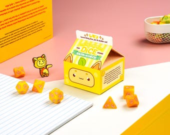 Könnte beinhalten: Ein gelbes Box-Set mit dem Text "Milk Carton Dice Games" und einem Cartoon-Zitronen-Charakter. Das Set enthält Würfel, einen Anstecker und Aufkleber. Mehrere gelbe Würfel sind um die Box herum verstreut, mit einem rosa und weißen Hintergrund.