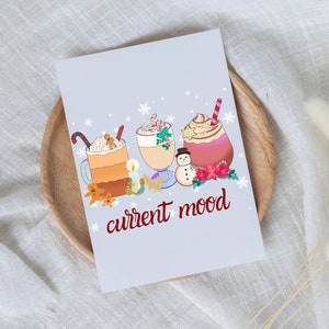 May include: A light blue card with a wintery scene of three mugs of hot chocolate with whipped cream and toppings. The text "current mood" is written in red below the mugs.