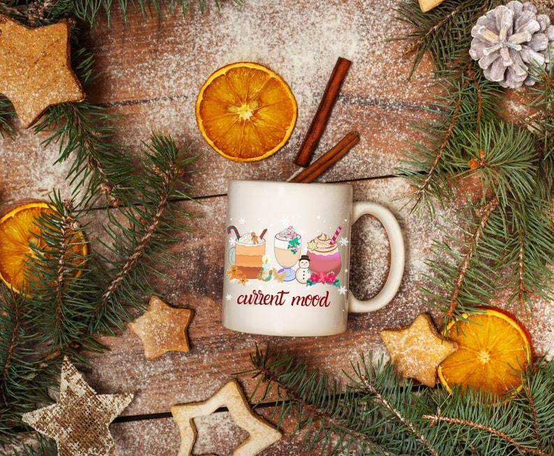 May include: White ceramic mug with a winter-themed design featuring three mugs of hot beverages with whipped cream and toppings. The mug has the text "current mood" printed on the front.