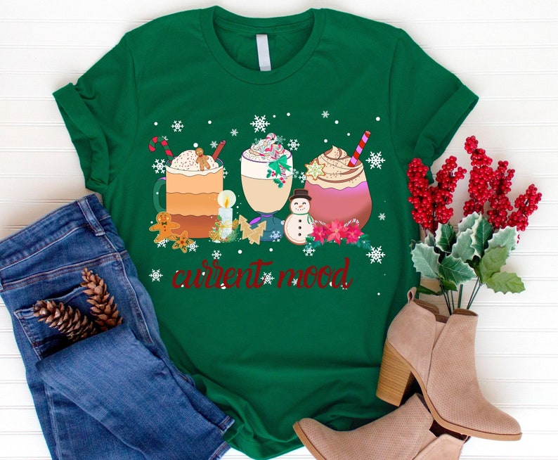 May include: Green t-shirt with a Christmas-themed design featuring three mugs of hot chocolate with whipped cream and candy canes, a snowman, and the text "current mood".
