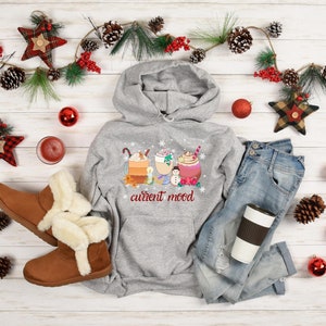 May include: A gray hoodie with a graphic of three holiday-themed drinks and the text "current mood". The hoodie is laid out on a white wood background with a pair of brown boots, a pair of blue jeans, a red and white plaid star, and a red and white plaid ornament.