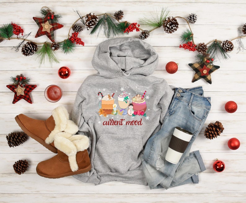 May include: A gray hoodie with a graphic of three holiday-themed drinks and the text "current mood". The hoodie is laid out on a white wood background with a pair of brown boots, a pair of blue jeans, a red and white plaid star, and a red and white plaid ornament.