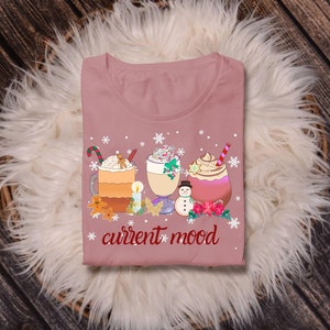 May include: A pink t-shirt with a winter-themed design featuring three mugs of hot beverages, a snowman, and the text "current mood".