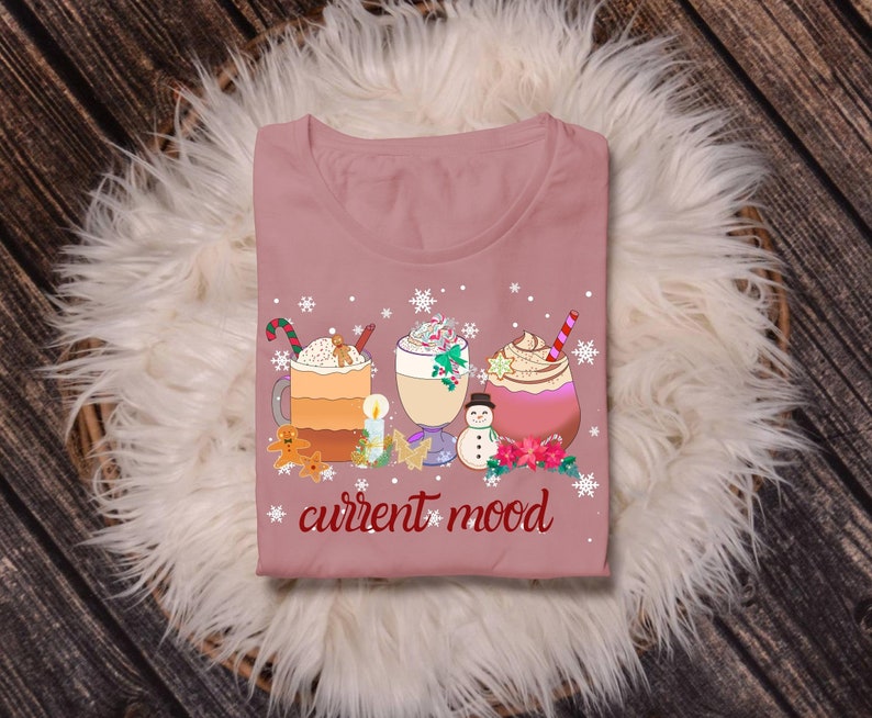May include: A pink t-shirt with a winter-themed design featuring three mugs of hot beverages, a snowman, and the text "current mood".