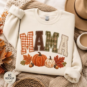 Peut inclure: Un sweat-shirt à col rond couleur crème portant le mot MAMA en grandes lettres majuscules. Chaque lettre présente un motif unique : vichy orange, velours côtelé marron, tartan vert et tricot torsadé crème. Citrouilles et feuilles d'automne.