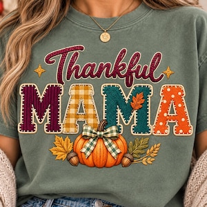Puede incluir: Una camiseta verde salvia con el texto Thankful MAMA en una fuente de estilo cosido con varios patrones. Debajo del texto hay una calabaza naranja con un lazo a cuadros verdes, rodeada de hojas de otoño y bellotas en un diseño gráfico.