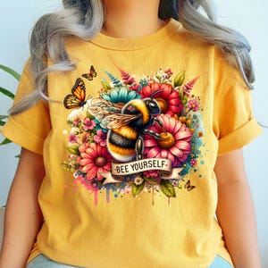 Pode incluir: Camiseta amarela de manga curta com um design floral colorido e uma abelha com o texto "BEE YOURSELF" em uma faixa.