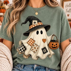 May include: A sage green t-shirt featuring a graphic of a ghost wearing a black witch hat with a plaid band. The ghost is decorated with various fabric patches in plaid and floral patterns, a small heart, and carries a jack-o'-lantern bucket.