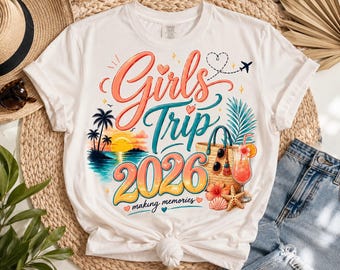 May include: A white t-shirt featuring a colorful graphic with the text Girls Trip 2026 making memories. The design includes a sunset over the ocean with palm trees, a straw tote bag, a tropical cocktail, a starfish, and a small airplane icon.