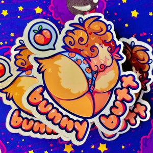 Könnte beinhalten: Ein Set mit drei Aufklebern, die einen Comic-Charakter mit pfirsichfarbenem Fell zeigen, der ein blaues und weiß gemustertes Outfit mit Pfirsichen trägt. Die Aufkleber haben den Text "Bummy Butt" in einer blasenartigen Schrift.