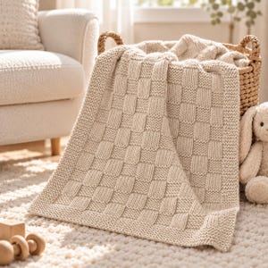 May include: A beige knitted blanket featuring a basketweave pattern with alternating squares of stockinette and garter stitches. The blanket is draped over a woven wicker basket, which also contains a small plush toy with long ears.