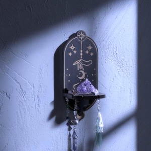 May include: A black wall shelf with celestial design elements, including stars, a hand, and a crescent moon. The shelf holds a cluster of purple crystals and dangling charms. The background is a textured, light blue wall.