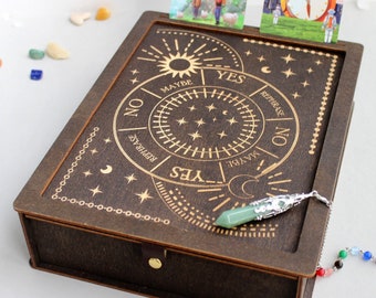 May include: A wooden box with a pendulum inside. The box has a circular design with the words "YES", "NO", "MAYBE", and "REPHRASE" around the edge. The pendulum is made of a green stone and a silver chain.