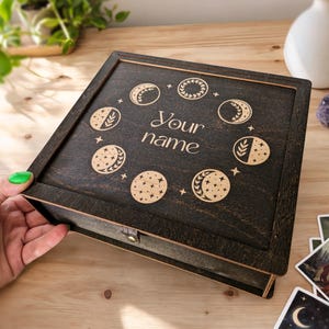 Puede incluir: Una caja de madera de color marrón oscuro con tapa abatible que presenta un diseño grabado con láser de fases lunares y estrellas dispuestas en círculo. El centro de la tapa muestra el texto Your name en una fuente cursiva.