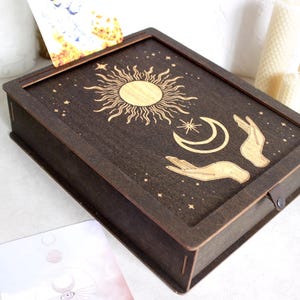 May include: A dark brown wooden box with a hinged lid. The lid features an intricate design of a sun, moon, and two hands holding the celestial bodies. The design is in a gold color.