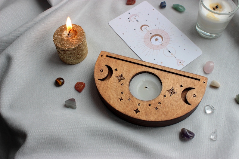May include: Wooden candle holder with a crescent moon design and a small candle in the center. The holder is surrounded by crystals and a card with a celestial design.