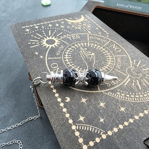 May include: A silver-toned metal pendulum featuring two smooth black spherical beads and a central decorative spacer with a small clear stone. The pendulum is attached to a silver chain and rests on a dark wooden board with gold celestial engravings.