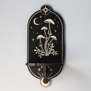 May include: Black wooden wall shelf with a crescent moon, stars, and mushrooms in a gold design. The shelf has a small ledge for displaying items.