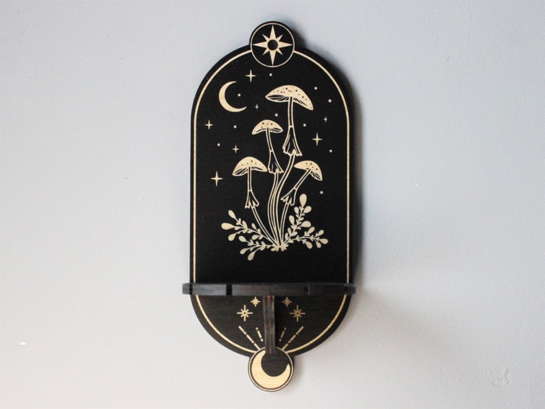 May include: Black wooden wall shelf with a crescent moon, stars, and mushrooms in a gold design. The shelf has a small ledge for displaying items.