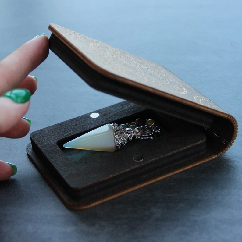 Puede incluir: Un p&eacute;ndulo de cristal blanco con una cadena de plata y una variedad de piedras de colores se muestra en una caja de madera con tapa abatible.