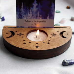 May include: Wooden candle holder with a crescent moon design and a small candle burning inside. The holder has a slot for a card, which reads "Believe in the impossible" and "Blue Moon".
