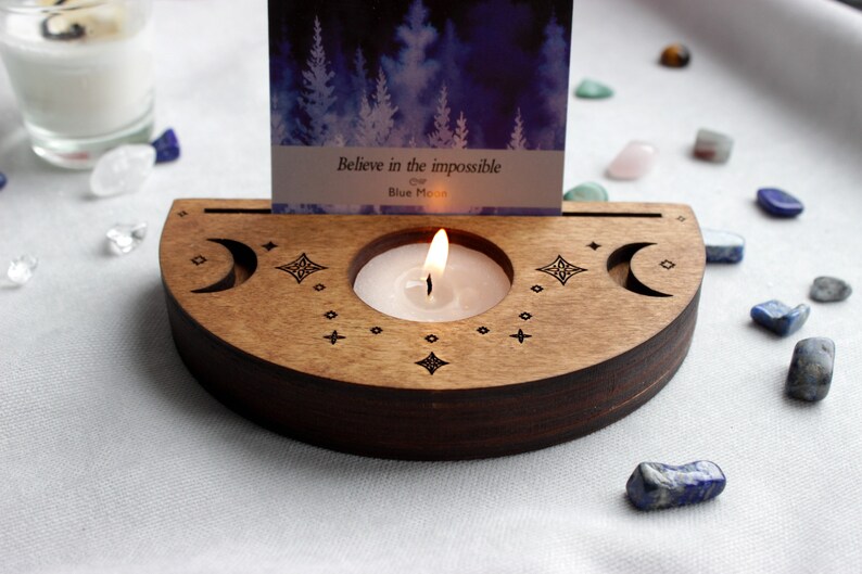 May include: Wooden candle holder with a crescent moon design and a small candle burning inside. The holder has a slot for a card, which reads "Believe in the impossible" and "Blue Moon".