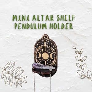 May include: Mini altar shelf pendulum holder made of dark wood, featuring a crescent moon design and a small shelf. The shelf holds a purple crystal and two silver chains. The text "MINI ALTAR SHELF PENDULUM HOLDER" is at the top.