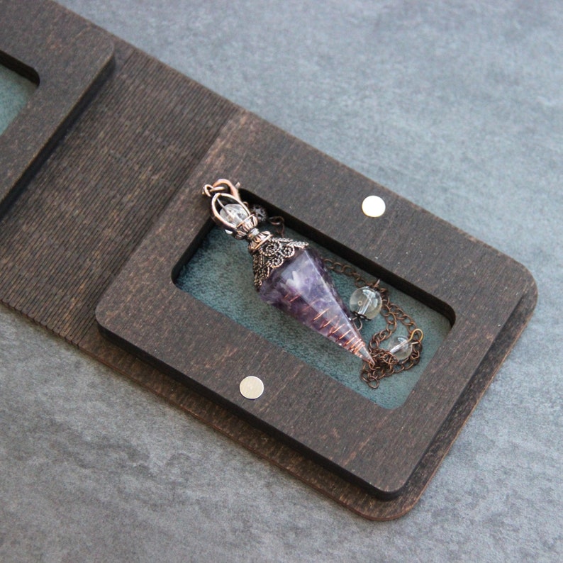 Puede incluir: Un p&eacute;ndulo de amatista p&uacute;rpura con un alambre de cobre envuelto alrededor de la punta, colgando de una cadena con cuentas transparentes, dentro de una caja de madera oscura con un forro de terciopelo.