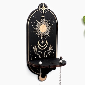 May include: Black wooden wall shelf with a sun, moon, and stars design. The shelf has two hooks and a small shelf for holding jewelry or other small items.