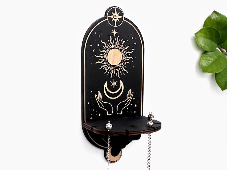 May include: Black wooden wall shelf with a sun, moon, and stars design. The shelf has two hooks and a small shelf for holding jewelry or other small items.