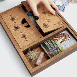 May include: A wooden box with a hinged lid, divided into compartments. The box contains a small plant, a crystal, and a deck of cards with a giraffe illustration. The box is decorated with intricate laser-cut designs.