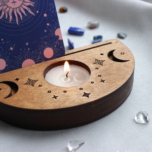 May include: A wooden candle holder with a crescent moon design and a small candle burning inside. The holder is decorated with stars and a small card with a sun and moon design is leaning against it.