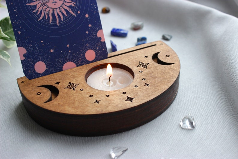 May include: A wooden candle holder with a crescent moon design and a small candle burning inside. The holder is decorated with stars and a small card with a sun and moon design is leaning against it.