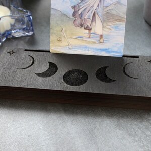 May include: A dark stained wooden card holder with moon phase cutouts. The holder displays a tarot card with a watercolor illustration. A star and crescent moon are carved into the wood. The holder is designed to display cards.