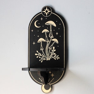 May include: Black wooden wall shelf with a crescent moon and star design. The shelf features a cutout of mushrooms and foliage in a gold color.