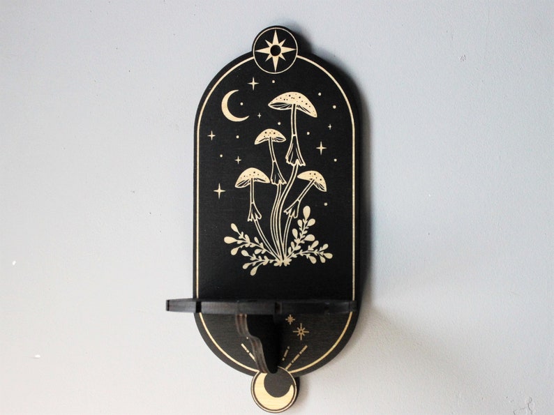May include: Black wooden wall shelf with a crescent moon and star design. The shelf features a cutout of mushrooms and foliage in a gold color.