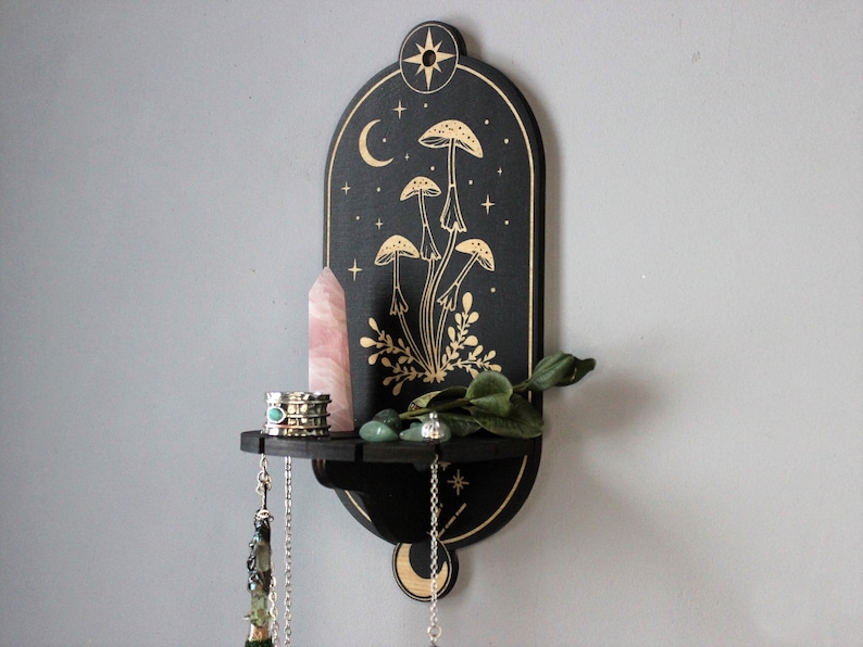 May include: Black wooden wall shelf with a crescent moon and stars design. The shelf features a gold mushroom illustration and a small shelf for displaying items.